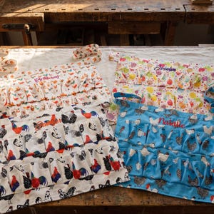 May include: Egg gathering aprons in various patterns. One is white with colorful roosters, another is blue with white chickens, and a third is white with colorful butterflies and chickens. Each apron has multiple pockets for collecting eggs.