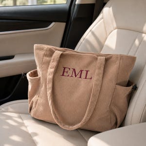 May include: A tan corduroy tote bag featuring two shoulder straps and side pockets with black drawstring toggles. The front of the bag is embroidered with the letters EML in a dark maroon serif font, centered on the textured fabric surface.