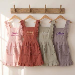 May include: Four ruffled aprons in pink, red, green, and purple hang on a wooden rack. Each apron has a different name embroidered on the front: Jennifer, Emily, Ashley, and Megan. The aprons have pockets and are striped.