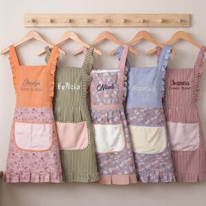 May include: Five personalized aprons hanging on wooden hangers. Each apron features ruffles on the straps and hem, a front pocket, and embroidered names including Jocelyn, Felicia, Nicole, Amanda, and Joanna. The aprons display various patterns like floral prints and vertical stripes in orange, green, purple, blue, and red.