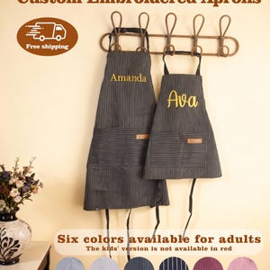 May include: Two black and white pinstriped aprons with black neck straps and waist ties. The larger apron features the embroidered name Amanda in yellow, and the smaller apron features the embroidered name Ava in yellow. Both aprons have a front pocket.