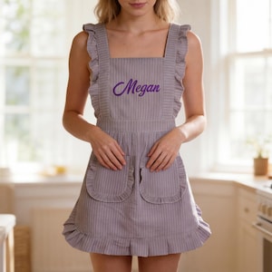 K&ouml;nnte beinhalten: Eine hellviolette Sch&uuml;rze mit d&uuml;nnen wei&szlig;en vertikalen Streifen, breiten ger&uuml;schten Schultertr&auml;gern, einem eckigen Ausschnitt und zwei Vordertaschen mit ger&uuml;schten R&auml;ndern. Der Name Megan ist in violetter Schreibschrift auf der Brust gestickt.