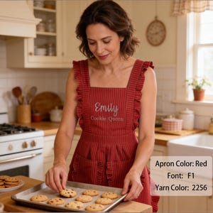 K&ouml;nnte beinhalten: Eine rote Sch&uuml;rze mit d&uuml;nnen wei&szlig;en vertikalen Streifen, ausgestattet mit ger&uuml;schten Schultertr&auml;gern und zwei Vordertaschen. Im Brustbereich ist der Name Emily und der Text Cookie Queen mit hellem Garn mittig auf den Stoff gestickt.