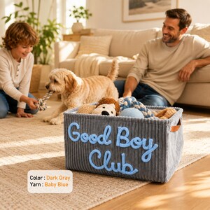May include: A rectangular dark gray corduroy storage basket featuring the text Good Boy Club in raised baby blue yarn lettering. The basket contains various plush toys and rope items, with brown leather-like side handles for easy carrying.