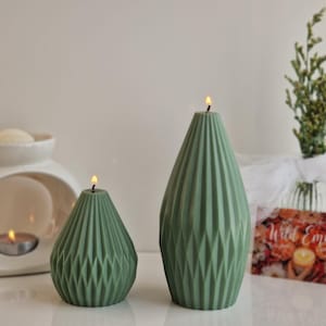 May include: Two sage green candles with a geometric, faceted, diamond-like texture. The candles are teardrop-shaped and feature a lit wick at the top. The smaller candle is on the left, and the taller, more elongated candle is on the right.