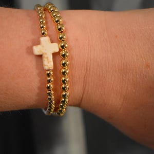 May include: A double-strand stretch bracelet featuring small, reflective gold-toned metallic beads. One strand includes a single, off-white, speckled cross-shaped bead as a central focal point, while the other strand consists entirely of gold beads.