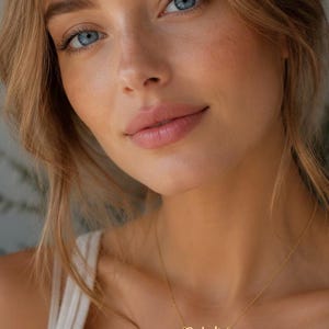 Könnte beinhalten: Eine goldfarbene Halskette mit dem Namen "Ophelia" in Schreibschrift. Die Halskette wird von einer Person mit heller Haut, blauen Augen und blondem Haar getragen. Die Person trägt ein weißes Tanktop.