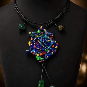 May include: A vibrant glass pendant necklace with an abstract design. The pendant features blue, red, yellow, and green colours. The necklace has a black cord with green and black bead details. The pendant is displayed on a black mannequin.
