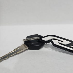 May include: A black and white key chain with a white "R3" logo. The keychain is attached to a silver key ring and a black key.