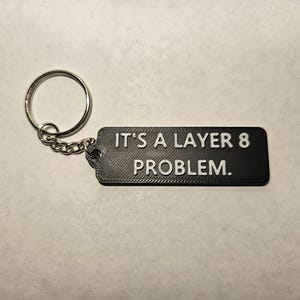 May include: Black rectangular keychain with the text "IT'S A LAYER 8 PROBLEM." in white. The keychain is attached to a silver key ring with a small chain.