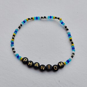Könnte beinhalten: Ein blaues, weißes und gelbes Perlenarmband mit dem Wort "PHANTOM" in schwarzen Perlen.