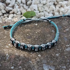 Könnte beinhalten: Ein handgefertigtes Armband mit dem Namen "MERCEDES" in weißen Buchstaben auf schwarzen quadratischen Perlen. Das Armband hat ein geflochtenes Design mit türkisfarbenen und grauen Fäden und wird auf einer strukturierten Oberfläche präsentiert.