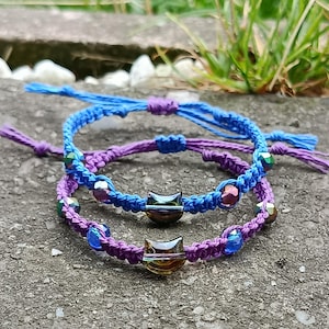 Puede incluir: Dos pulseras de macramé hechas a mano en azul y morado. Cada pulsera presenta cuentas iridiscentes y un dije en forma de gato. Las pulseras están anudadas y son ajustables. Se muestran sobre una superficie gris.
