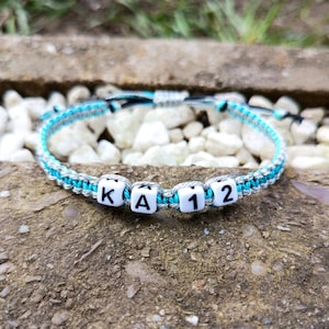Peut inclure: Bracelet fait main avec un design tressé gris et turquoise. Le bracelet comporte des perles carrées blanches avec les lettres "K", "A" et les chiffres "12". Le bracelet est présenté sur une surface rugueuse et texturée.
