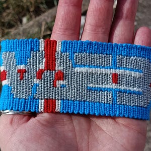 Pode incluir: Pulseira tecida azul, vermelha e branca. A pulseira apresenta um design geométrico com fundo azul, detalhes vermelhos e detalhes brancos e cinzentos. A pulseira tem cerca de 4 cm de largura.