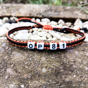 Puede incluir: Pulsera hecha a mano con cord&oacute;n trenzado negro y naranja. Las cuentas cuadradas blancas deletrean "OP81". La pulsera se muestra sobre una superficie rugosa y texturizada, con peque&ntilde;as piedras blancas y vegetaci&oacute;n en el fondo.