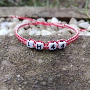 Peut inclure: Un bracelet fait main avec un cordon rouge et blanc tressé. Le bracelet comporte des perles carrées blanches avec les lettres "L", "H" et les chiffres "44" en noir. Le bracelet repose sur une surface grise texturée.