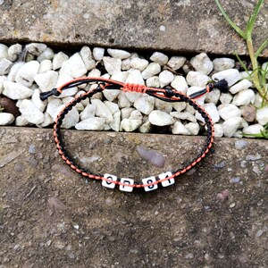 Puede incluir: Pulsera trenzada negra y naranja con cuentas blancas con letras "CP 81". La pulsera est&aacute; sobre un fondo de guijarros blancos y hormig&oacute;n. Art&iacute;culo hecho a mano.