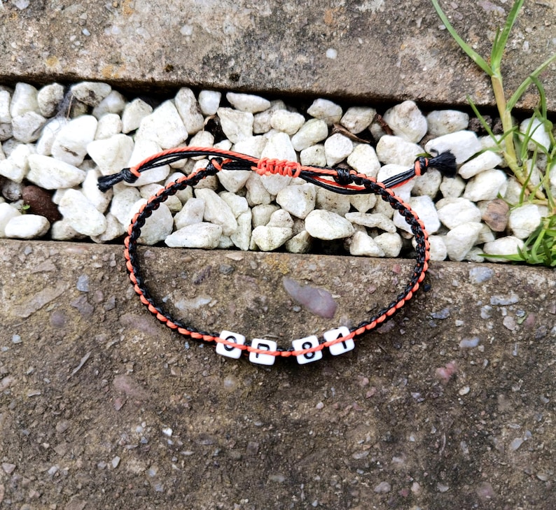 Puede incluir: Pulsera trenzada negra y naranja con cuentas blancas con letras "CP 81". La pulsera est&aacute; sobre un fondo de guijarros blancos y hormig&oacute;n. Art&iacute;culo hecho a mano.
