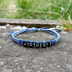 Puede incluir: Una pulsera hecha a mano con el nombre "JOOST" escrito con cuentas de letras negras y blancas. La pulsera presenta un diseño tejido azul y blanco y se muestra sobre una superficie gris.