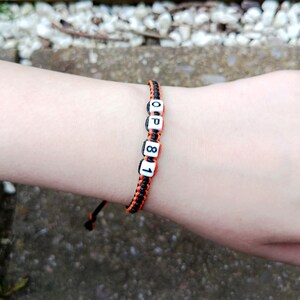 Puede incluir: Pulsera trenzada negra y naranja con cuentas blancas con letras "OP81". La pulsera est&aacute; en una mu&ntilde;eca, con un fondo borroso de una superficie de piedra.