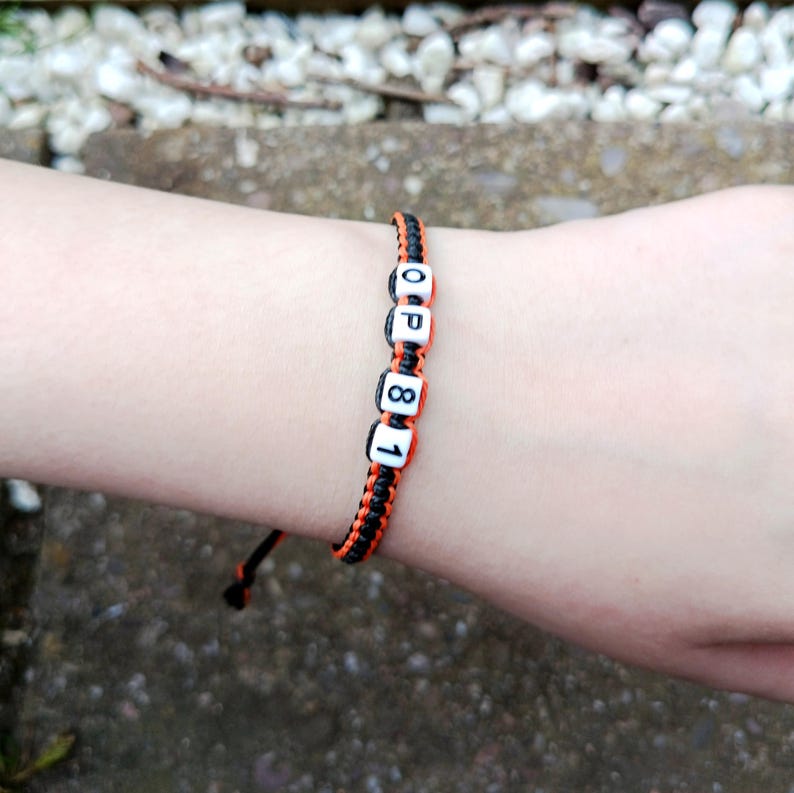 Puede incluir: Pulsera trenzada negra y naranja con cuentas blancas con letras "OP81". La pulsera est&aacute; en una mu&ntilde;eca, con un fondo borroso de una superficie de piedra.