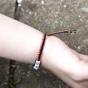 Puede incluir: Una pulsera de macram&eacute; negra y naranja con cuentas de letras blancas que deletrean "OF". La pulsera se lleva en la mu&ntilde;eca y presenta un dise&ntilde;o trenzado con hilos naranjas y negros. La pulsera est&aacute; atada con un nudo.
