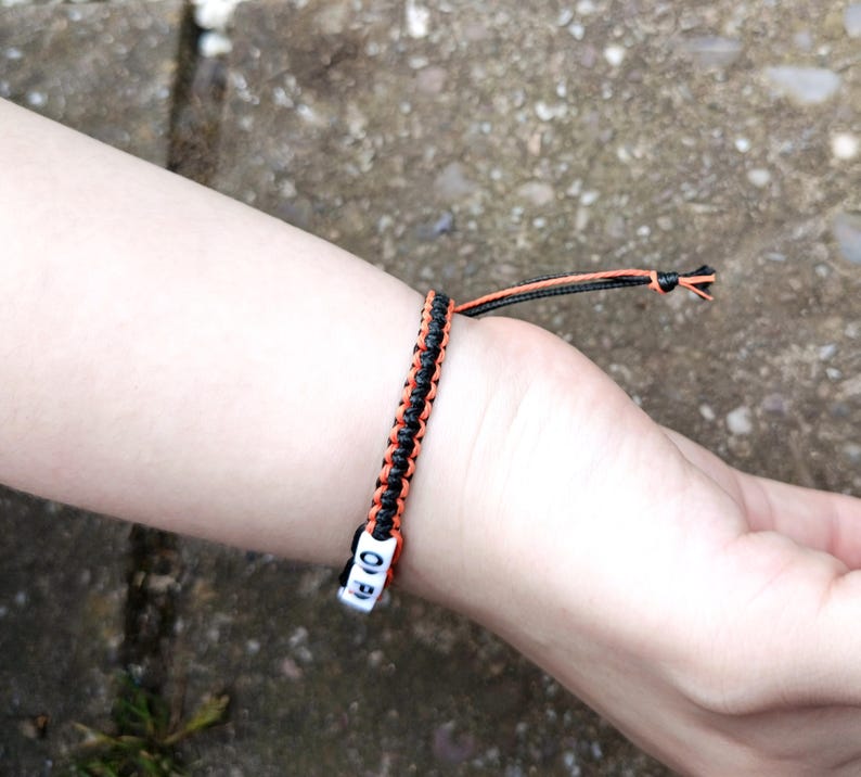 Puede incluir: Una pulsera de macram&eacute; negra y naranja con cuentas de letras blancas que deletrean "OF". La pulsera se lleva en la mu&ntilde;eca y presenta un dise&ntilde;o trenzado con hilos naranjas y negros. La pulsera est&aacute; atada con un nudo.