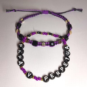 Puede incluir: Dos pulseras de cuentas moradas con cuentas negras y detalles dorados. Una pulsera deletrea "PAPA" y la otra "PERPETUA".