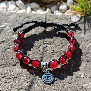 May include: A handmade bracelet featuring red beads and a black woven cord. The bracelet has a silver charm with the Cancer zodiac symbol. The beads are a mix of solid and faceted, creating a textured look.