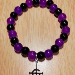 Op de afbeelding: Een armband met afwisselend zwarte en paarse kralen. Een zilverkleurige bedel met een kruisontwerp hangt aan de armband. De armband wordt getoond op een lichtbruin houten oppervlak.