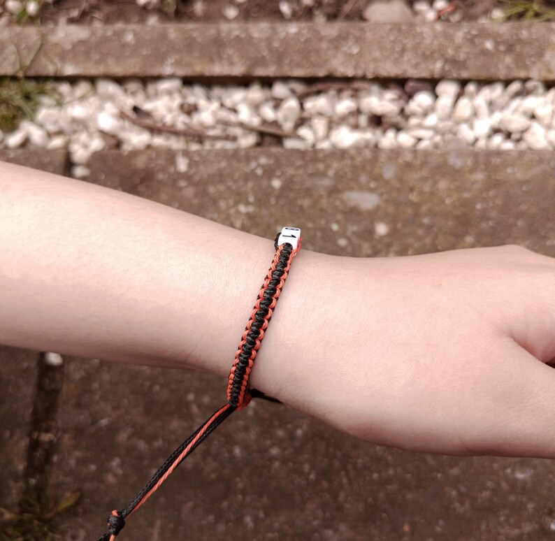 Puede incluir: Una pulsera trenzada negra y naranja con un cierre plateado. La pulsera est&aacute; hecha de cord&oacute;n tejido y se lleva en la mu&ntilde;eca. El fondo est&aacute; desenfocado.