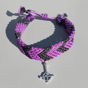 Op de afbeelding: Zwart en paars geweven armband met een chevronpatroon. De armband heeft een zilverkleurige kruisbedel. Het is verstelbaar met een schuifknoopsluiting. De armband is gemaakt met macramé.