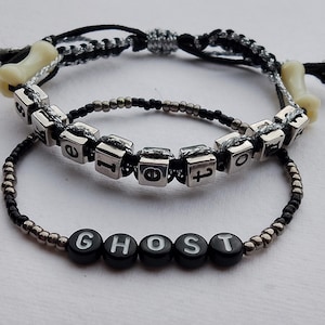 Op de afbeelding: Twee armbanden met zwarte kralen en zilveren bedels. De bovenste armband heeft het woord "geloven" gespeld in zilveren bedels. De onderste armband heeft het woord "geest" gespeld in zwarte kralen.
