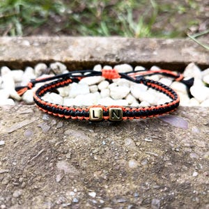 Peut inclure: Bracelet en macramé noir et orange avec des perles dorées portant les lettres "L" et "N". Le bracelet est posé sur une surface rugueuse et texturée, avec de petits cailloux blancs en arrière-plan.