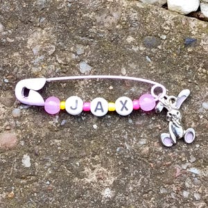Könnte beinhalten: Eine hellviolette Sicherheitsnadel mit drei weißen runden Perlen, die die schwarzen Buchstaben J, A und X tragen. Die Nadel ist mit kleinen rosa und gelben Zwischenperlen sowie einem silberfarbenen Metallanhänger in Hasenform verziert.