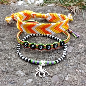 Può includere: Tre braccialetti fatti a mano. Il braccialetto superiore presenta un motivo a zig-zag arancione, giallo e bianco. Il braccialetto centrale ha la parola "SPOOKY" scritta con perline nere con lettere verdi. Il braccialetto inferiore ha un ciondolo a forma di ragno argentato.