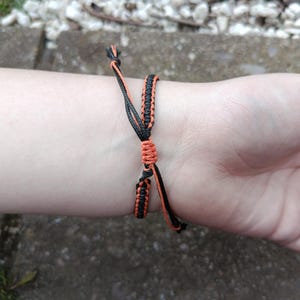 Puede incluir: Pulsera de macram&eacute; negra y naranja hecha a mano, atada alrededor de una mu&ntilde;eca. La pulsera presenta un dise&ntilde;o tejido con hilos naranjas y negros. La pulsera es ajustable. El fondo est&aacute; borroso.
