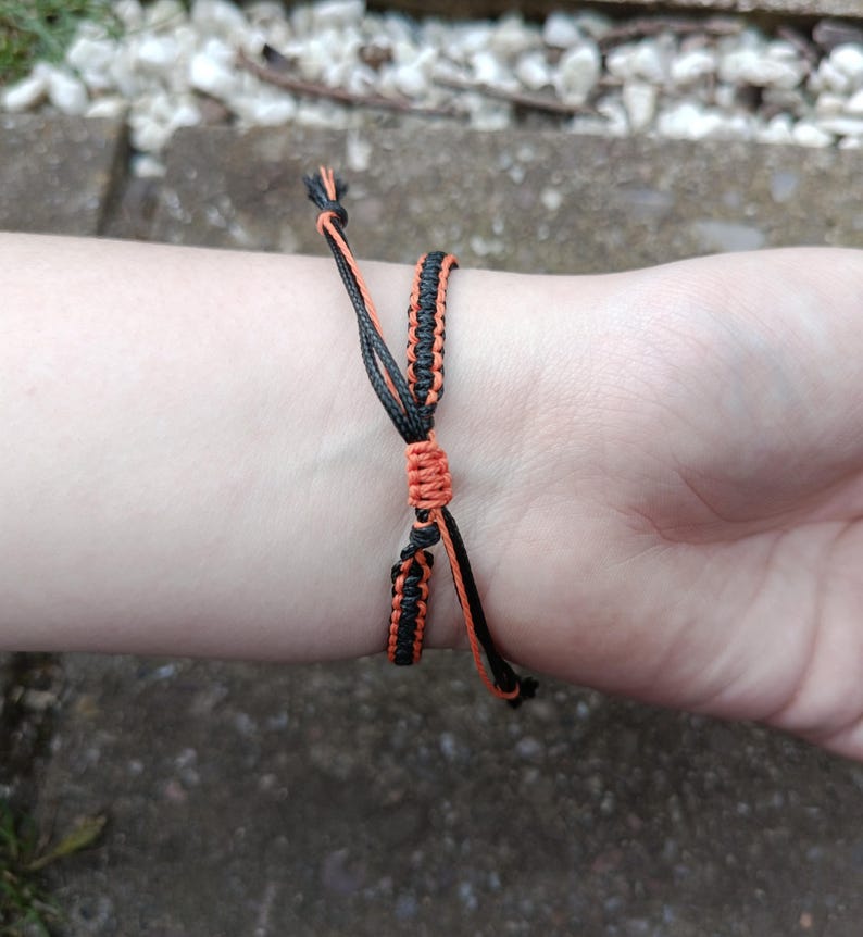 Puede incluir: Pulsera de macram&eacute; negra y naranja hecha a mano, atada alrededor de una mu&ntilde;eca. La pulsera presenta un dise&ntilde;o tejido con hilos naranjas y negros. La pulsera es ajustable. El fondo est&aacute; borroso.