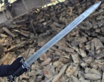 Op de afbeelding: Een lang, zilveren zwaard met een patroon op het lemmet en een zwarte handgreep. Het zwaard heeft een decoratieve pareerstang en een punt. De handgreep is omwikkeld met zwart materiaal en heeft zilveren accenten. Het zwaard wordt tegen een achtergrond van hout gehouden.