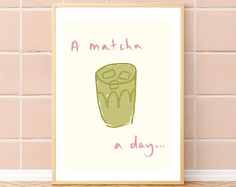 May include: A framed wall art print featuring a light cream background with a hand-drawn illustration of a green matcha drink in a glass with ice cubes. The pink text above and below the illustration reads A matcha a day.
