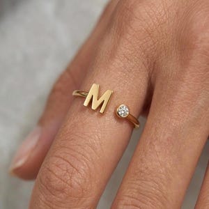 Peut inclure: Une bague dorée avec la lettre "M" et une petite pierre précieuse claire. La bague est portée au doigt, soulignant son design délicat et sa finition polie. La pierre précieuse est sertie dans un sertissage à lunette.