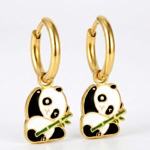 Peut inclure: Deux boucles d'oreilles dorées à créoles avec des breloques en forme de panda. Chaque breloque représente un panda blanc avec des détails noirs, tenant une tige de bambou verte. Fond blanc.
