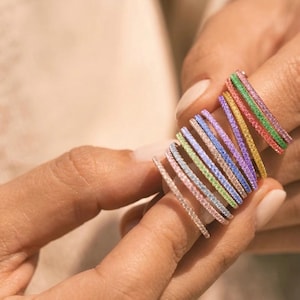 Op de afbeelding: Een stapel dunne, verfijnde ringen met een doorlopende rij kleine, pavé gezette edelstenen in een levendig regenboogspectrum. De collectie bevat individuele ringen in tinten roze, groen, geel, paars, blauw en lichtoranje.