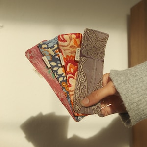 May include: A hand holds a fan of patterned fabric cases. The cases feature floral and paisley designs in red, blue, orange, and brown. Each case has a small white label with the name "Khalifa".