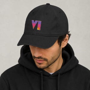 May include: A black baseball cap featuring an embroidered logo on the front. The logo consists of the letters VI in a stylized font, filled with a gradient pattern of purple, pink, and orange horizontal stripes. The cap has a curved brim.