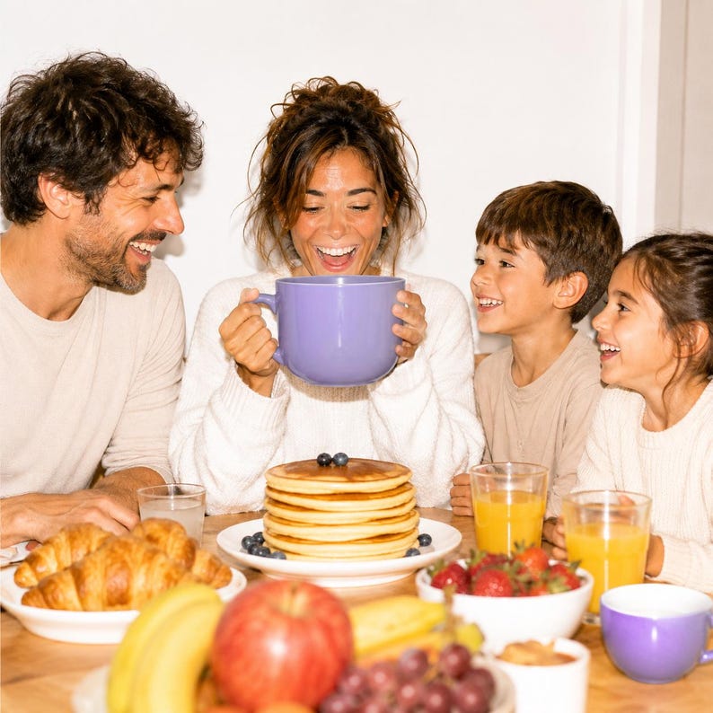 Peut inclure: Une grande tasse en c&eacute;ramique violet clair tenue par une personne, plac&eacute;e au-dessus d'une pile de cr&ecirc;pes dor&eacute;es garnies de myrtilles. La table pr&eacute;sente une assiette de croissants, un bol de fraises, des bananes, une pomme et du raisin.