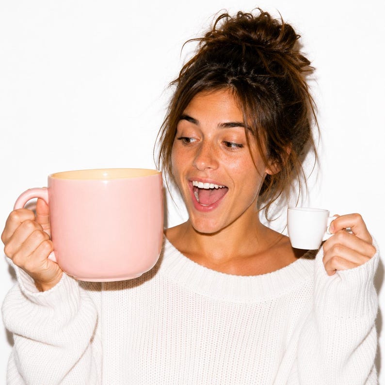 Peut inclure: Une grande tasse en c&eacute;ramique rose clair lisse avec une anse arrondie tenue dans une main, contrast&eacute;e par une minuscule tasse &agrave; expresso en c&eacute;ramique blanche unie tenue dans l'autre main, montrant une diff&eacute;rence d'&eacute;chelle entre les deux.