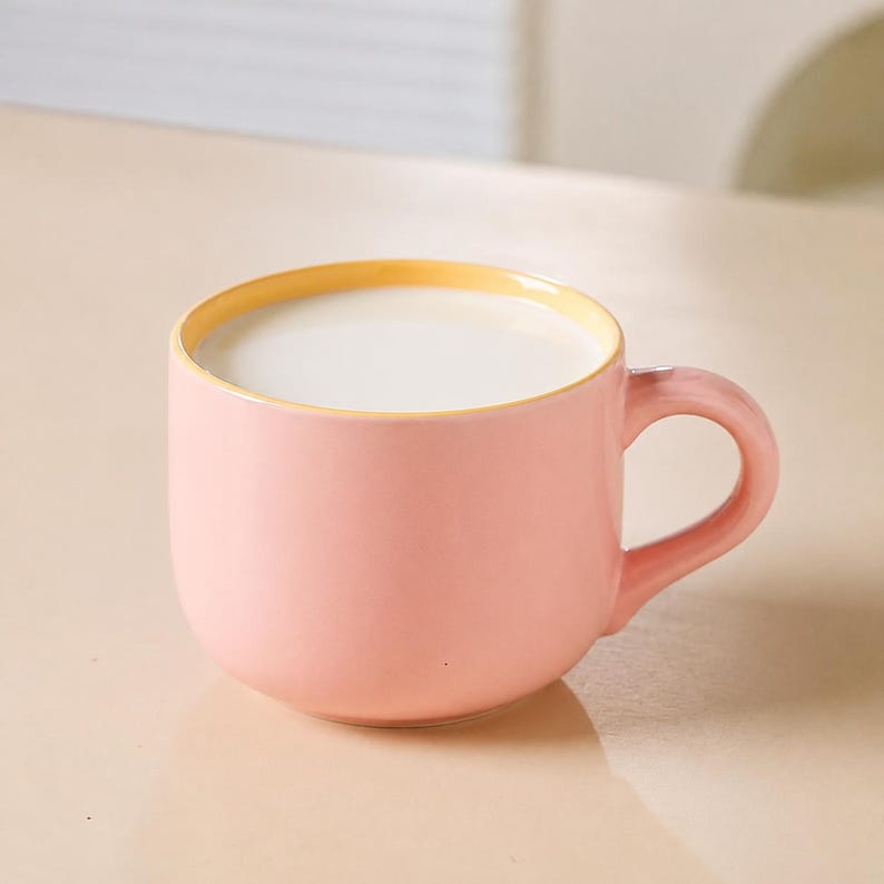 Peut inclure: Une tasse en c&eacute;ramique ronde avec un ext&eacute;rieur rose doux et un bord int&eacute;rieur jaune p&acirc;le. La tasse pr&eacute;sente une anse incurv&eacute;e sur le c&ocirc;t&eacute; droit et est remplie &agrave; ras bord d'un liquide blanc, cr&eacute;ant un aspect lisse et uniforme dans le r&eacute;cipient.