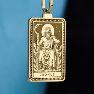 Puede incluir: Un collar con colgante rectangular de tono dorado que presenta una ilustración grabada de una figura barbuda sentada en un trono, sosteniendo una guadaña y un reloj de arena, con el texto CRONUS inscrito en la parte inferior, en una cadena de cuerda.